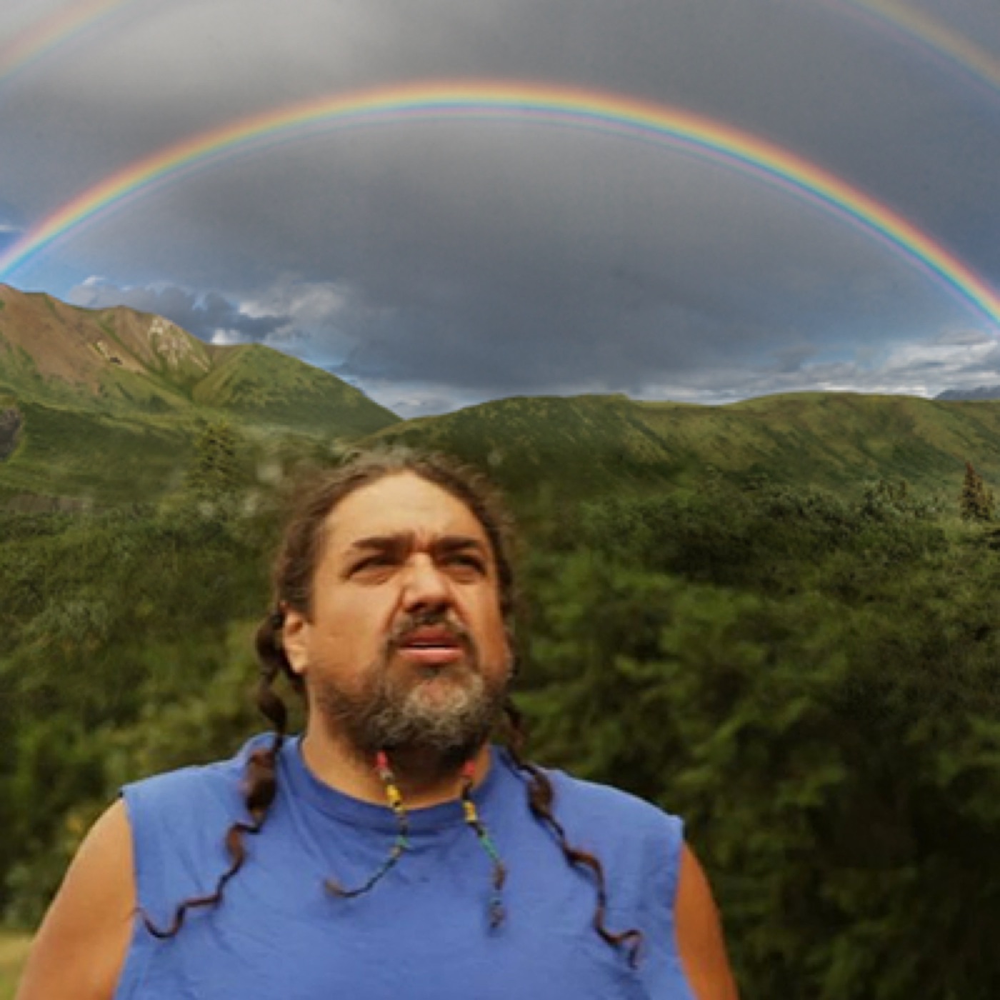 Bearded long-haired man in a blue tank top standing beneath a double rainbow over mountains