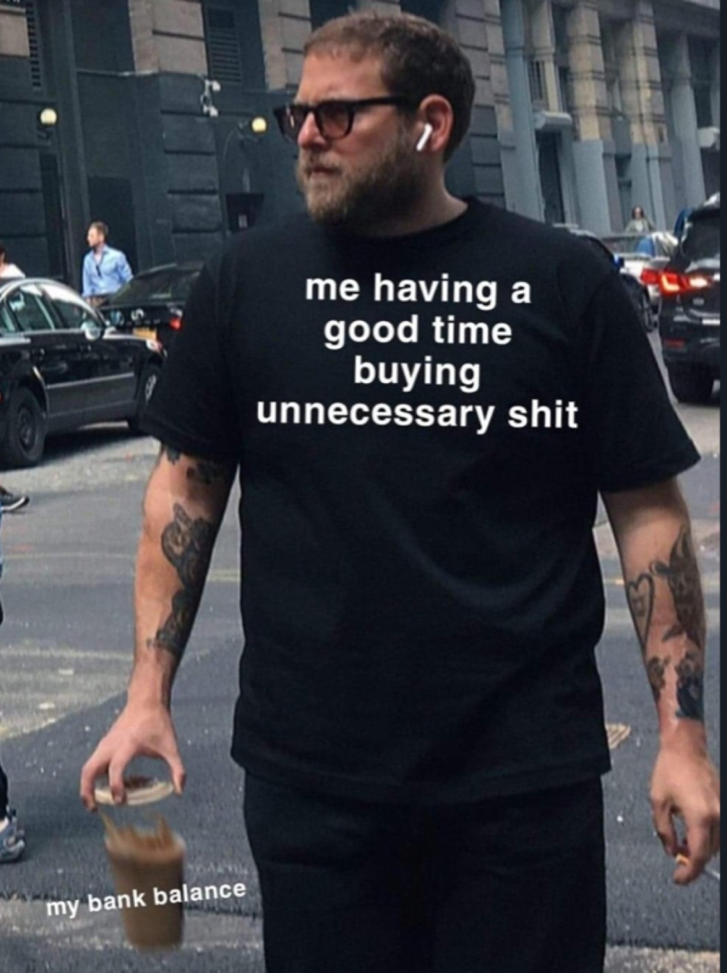 Bearded man in black t-shirt on a street fumbling an iced coffee, captioned "me having a good time buying unnecessary shit"