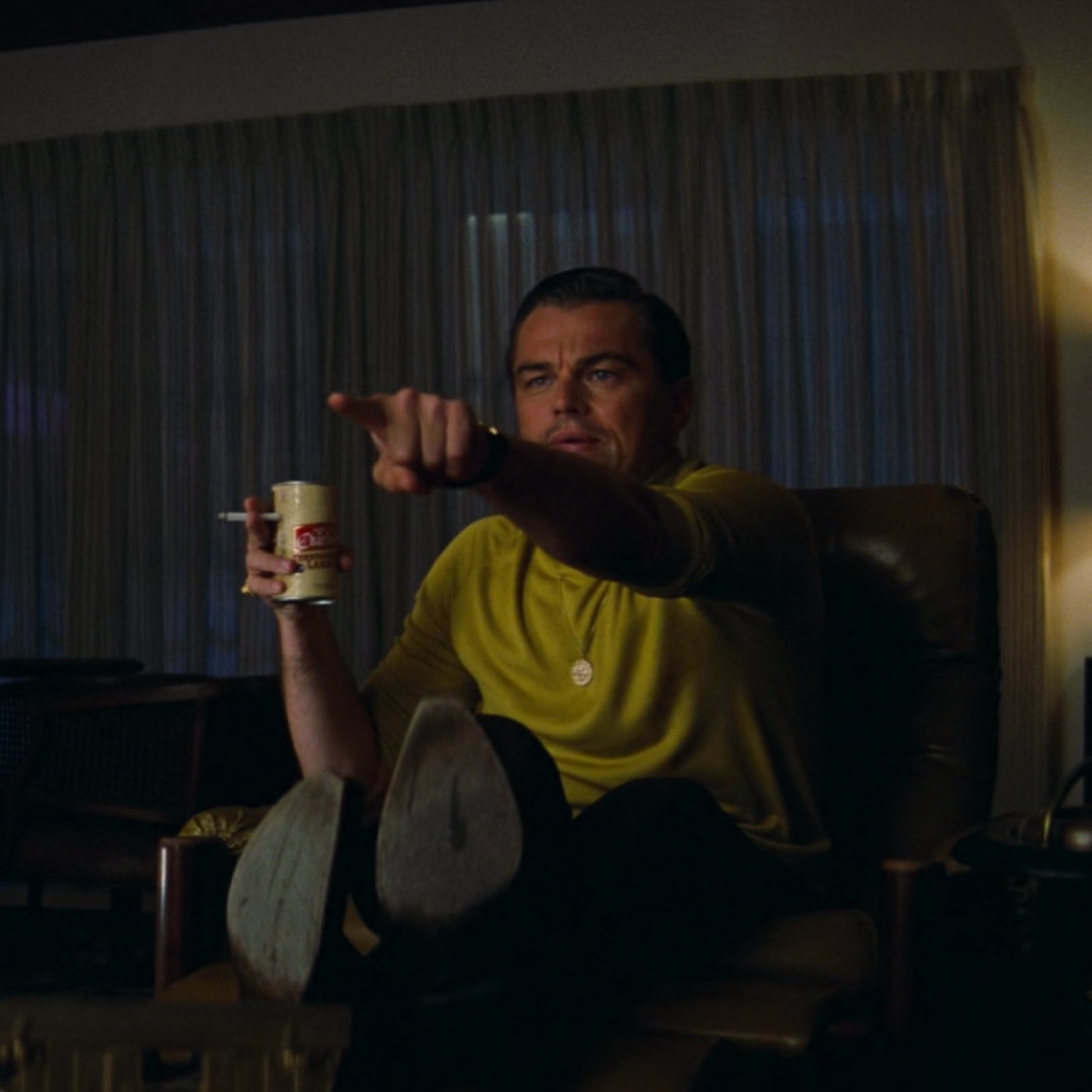 Man in a yellow shirt reclining with his feet up, beer can and cigarette in one hand, pointing excitedly toward the camera